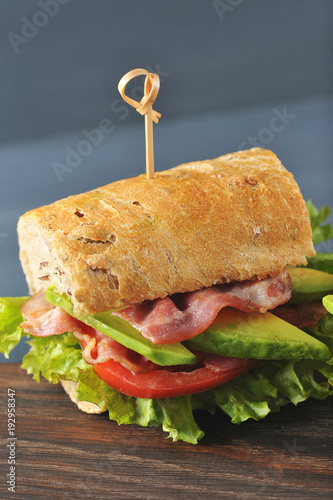 Appetizing sandwich on a wooden board. Baguette sandwich with filling from lettuce, slices of fried bacon, tomato and avocado. Dark background. Vertical orientation of the frame. Close-up. Macro photo