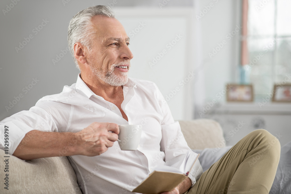 Coffee break. Attractive mature man keeping smile on his face and holding cup in right hand while having dreamy mood
