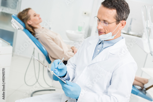 Dentist recording diagnosis while female patient waiting in modern clinic