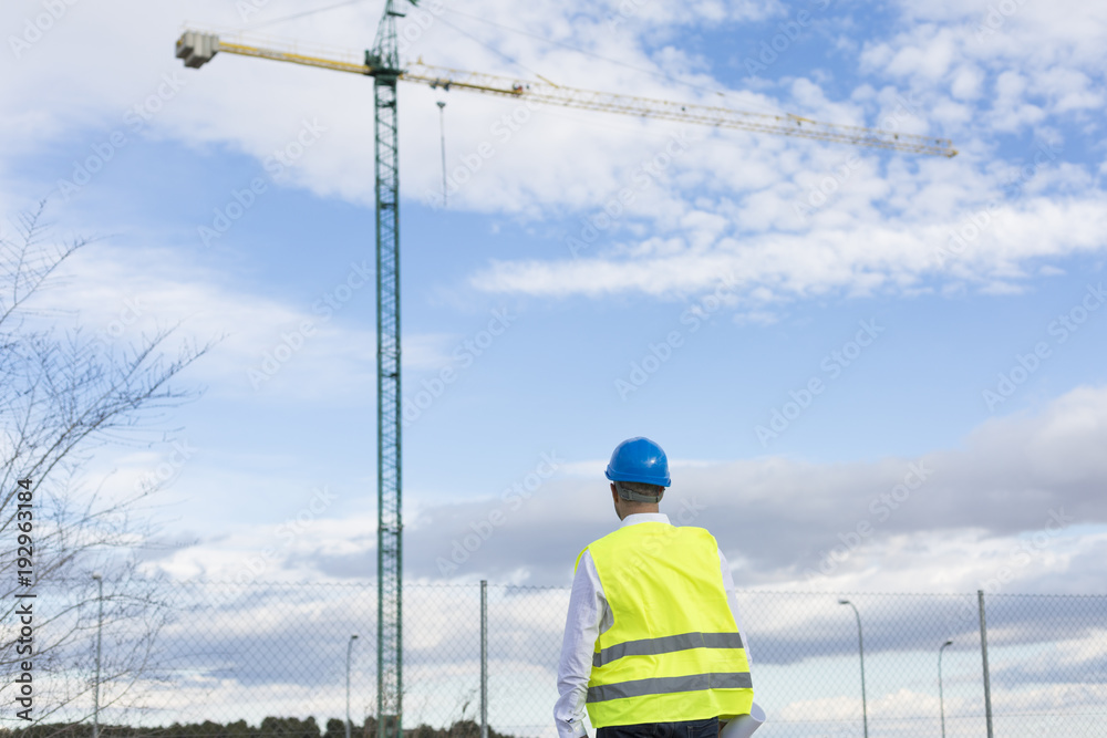 Architect or Engineer man holding blueprints on the Construction Site. Wearing protection clothes. Crane on background over blue sky. Job concept. Back light Architect or Engineer man holding blueprints on the Construction Site. Wearing protection clothes. Crane on background over blue sky. Job concept. Back light