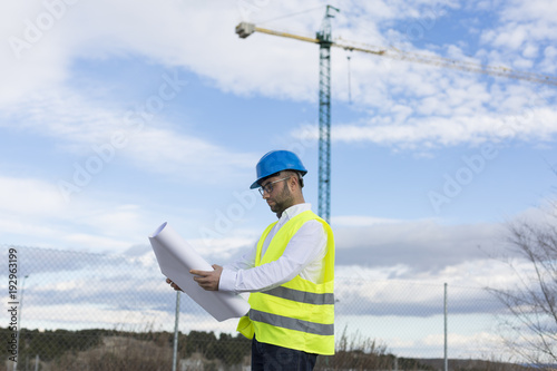 Architect or Engineer man holding blueprints on the Construction Site. Wearing protection clothes. Crane on background over blue sky. Job concept. Back light