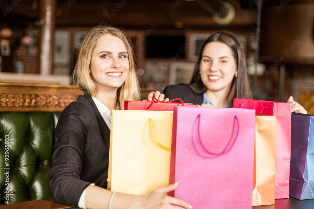 Young smiling blond and brunette at background watch at camera sitting in a cafe after shopping Young smiling blond and brunette at background watch at camera sitting in a cafe after shopping