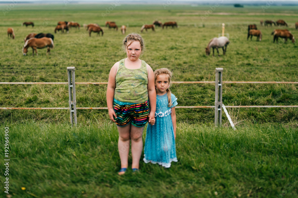Obraz premium Tilt shift blurred expressive mood potrtrait of two little sisters resting outdoor in summer together. Young girls with emotional faces. Chilhood in village. Funny kids near grazing horses field.