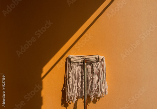 Old mop leaning against the orange wall have plenty of sunshine and shades.