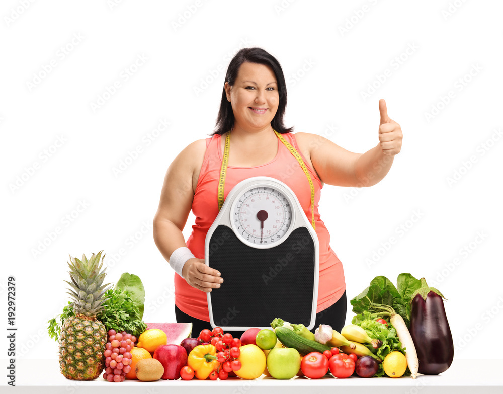 Overweight woman with a weight scale making a thumb up gesture behind a table with fruit and vegetables Overweight woman with a weight scale making a thumb up gesture behind a table with fruit and vegetables