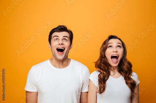 Ecstatic young man and woman in white t-shirts looking upward in excitement with open mouth, isolated over yellow background