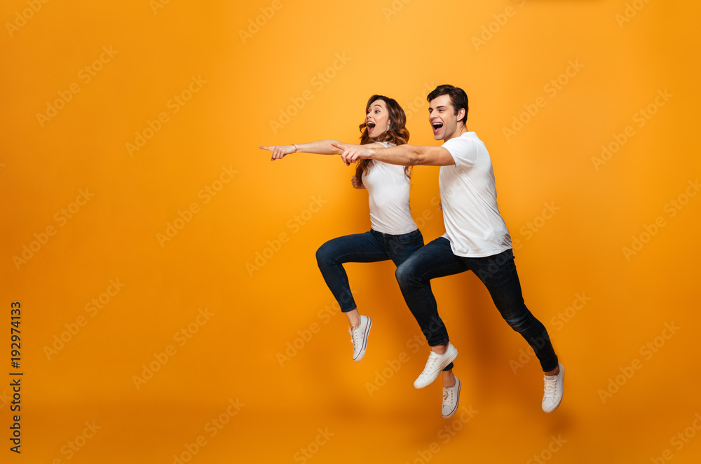 Photo of energetic beautiful couple posing together on camera while running or jumping and pointing fingers on copyspace, along background