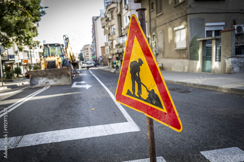 a sign of the road that shows that the road is in repair and workers are working on the street. the background is a yellow excavator. street repairs and a warning sign for asphalt and road repair.