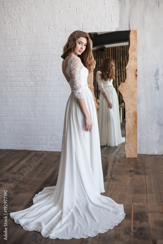 Beautiful bride looks in the mirror. Turned around. Floor-length dress. Dress with lace. A large mirror stands on the floor. White brick wall.