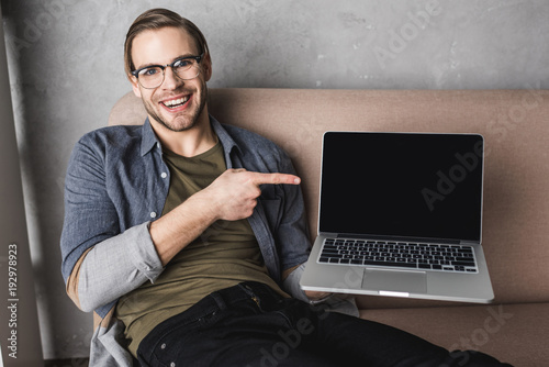 young smiling man sitting on couch and pointing at laptop screen