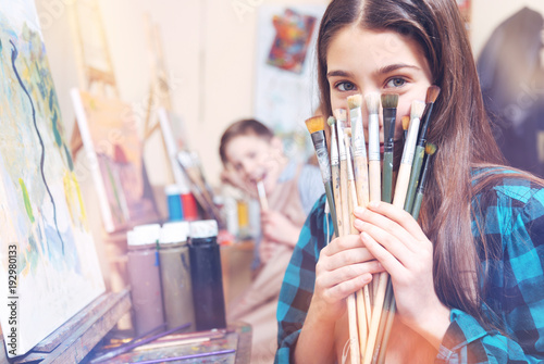 Creative studying. Pretty teenage girl hiding her face behind many painting brushes while sitting at a painting easel and smiling into the camera.