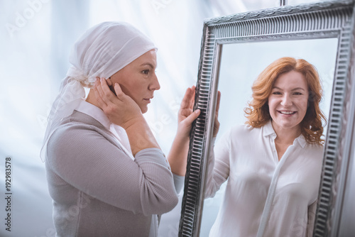 upset sick mature woman in kerchief looking at smiling reflection in mirror, cancer concept