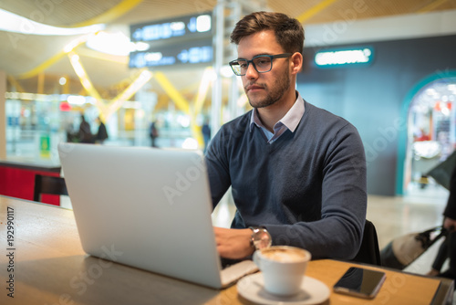 Fototapet young man at the airport working with laptop using his mobile phone and drinking