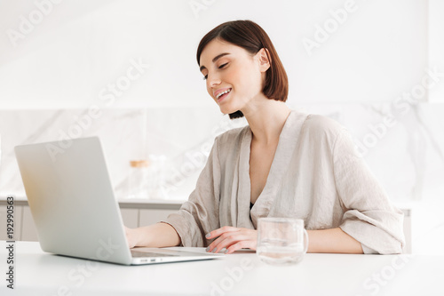 Portrait of caucasian adult woman wearing robe smiling and working, or chatting on white laptop in flat
