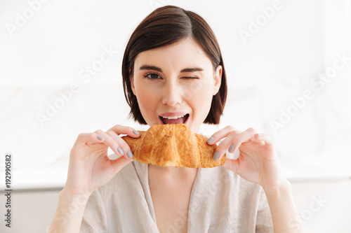 Image of happy young woman in robe having breakfast in hotel room, and eating tasty croissant in morning