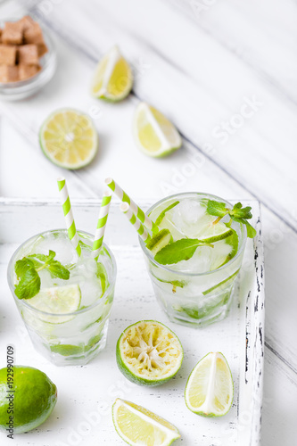 Caipirinha, mojito cocktail with lime, brown sugar, ice and mint leaves in beautiful glasses, cut green citrus on white wooden background. Summer alcohol drink. Close up photography. Selective focus