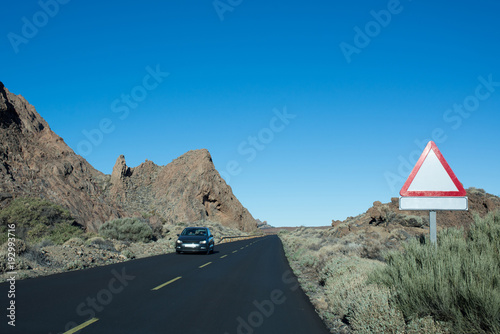 Highway goes through the rocks with green plants with empty road sign. Another car goes on your road on the another stripe.  Teide National Park. Tenerife