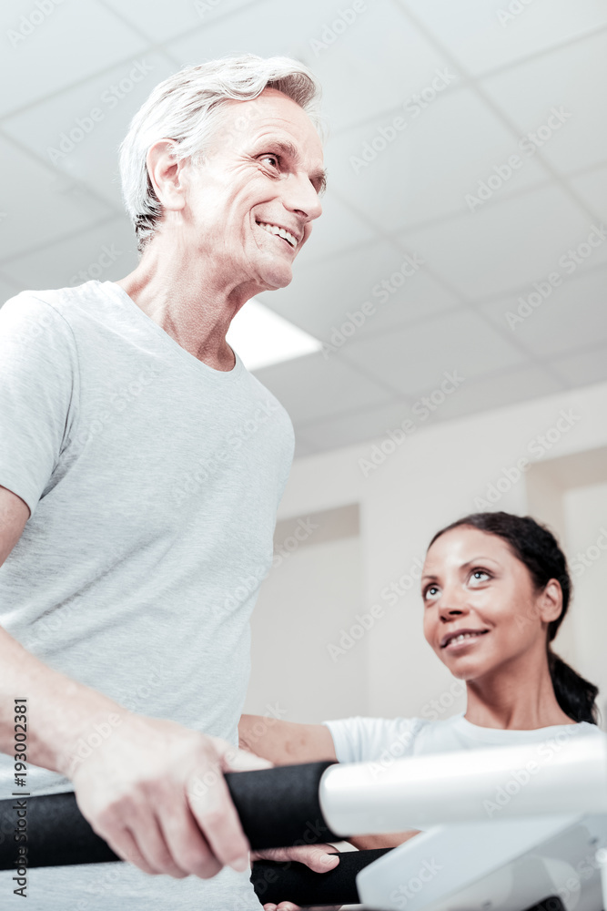 Recovery. Exuberant energetic old grey-haired man smiling and exercising on a training device and a smiling dark-haired dark-eyed woman sitting near him and looking at him Recovery. Exuberant energetic old grey-haired man smiling and exercising on a training device and a smiling dark-haired dark-eyed woman sitting near him and looking at him