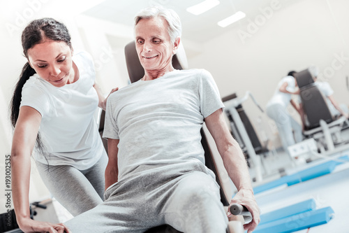Therapist. Gleeful determined old grey-haired man smiling and exercising on a training device while a dark-haired dark-eyed afro-american woman helping him