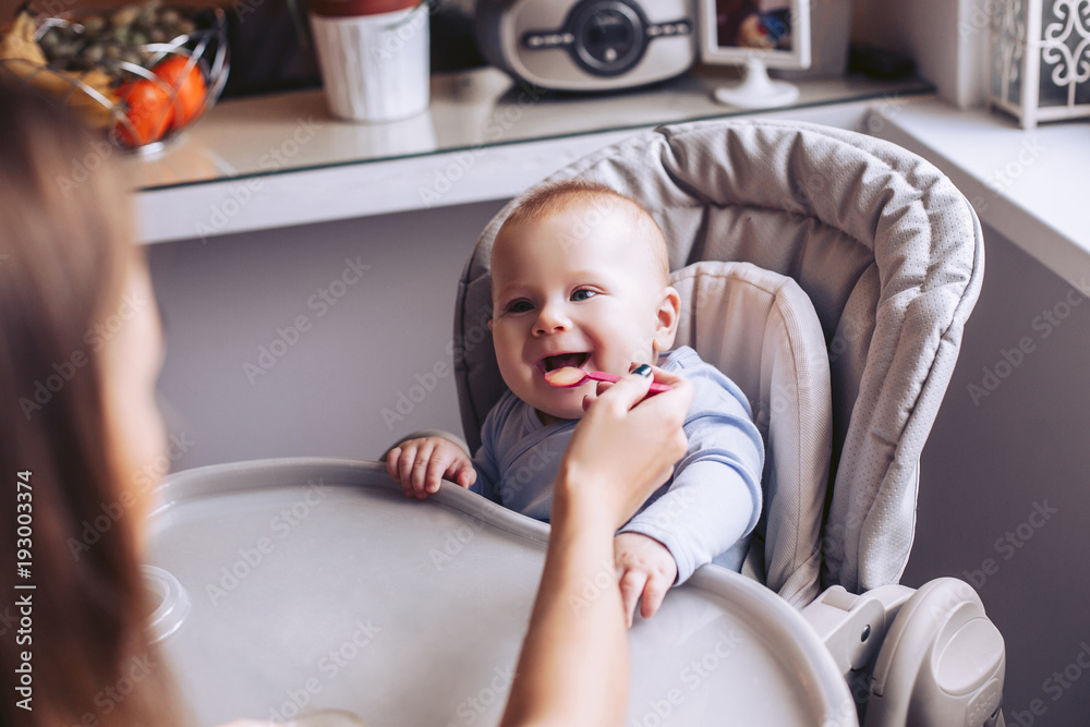 Mom feeds the baby from the spoon in the kitchen. A child is sitting in a child's chair. Mom feeds the baby from the spoon in the kitchen. A child is sitting in a child's chair.