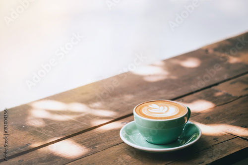 Hot cappuccino coffee in a green cup with latte art on the wooden table in a coffee shop and glass background. Vintage toned.