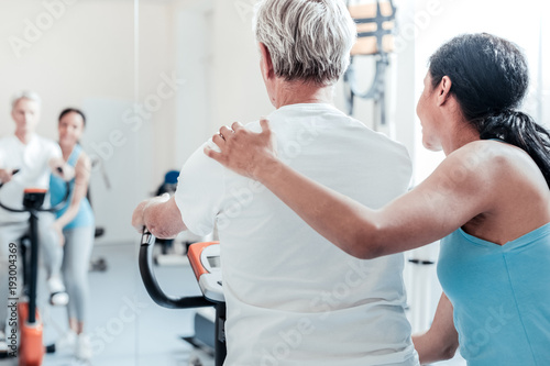 Exercising together. Old grey-haired man dressed in white shirt exercising on a training device while a young dark-haired afro-american female trainer standing near him and touching him on his back