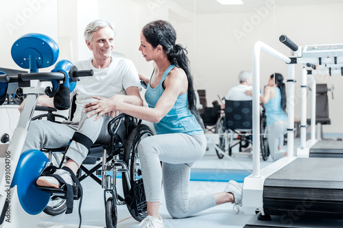 Helping others. Happy smiling old grey-haired man sitting in a wheelchair while a content young dark-haired female trainer smiling and standing near him and a training device in front of them