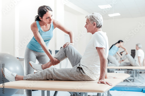 Stretching legs. Serious old grey-haired man sitting on bed while a concentrated young dark-haired afro-american female trainer stetching his legs and they looking at each other photo