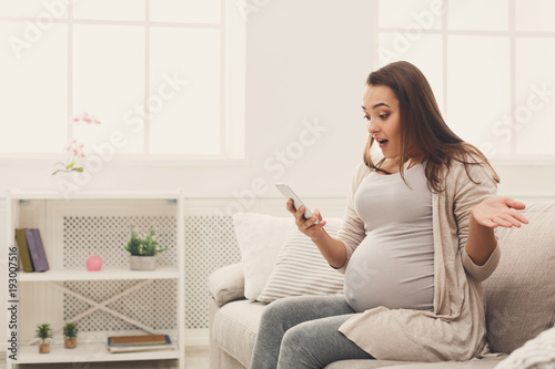 Surprised pregnant woman using smartphone at home