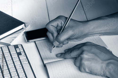 Hand on write on notebook with desktop keyboard, mobile phone in blue tone moode photo