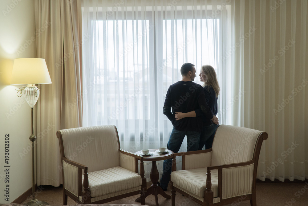 The newlyweds stand at the window of the hotel room and look at each other. Young family is very happy The newlyweds stand at the window of the hotel room and look at each other. Young family is very happy