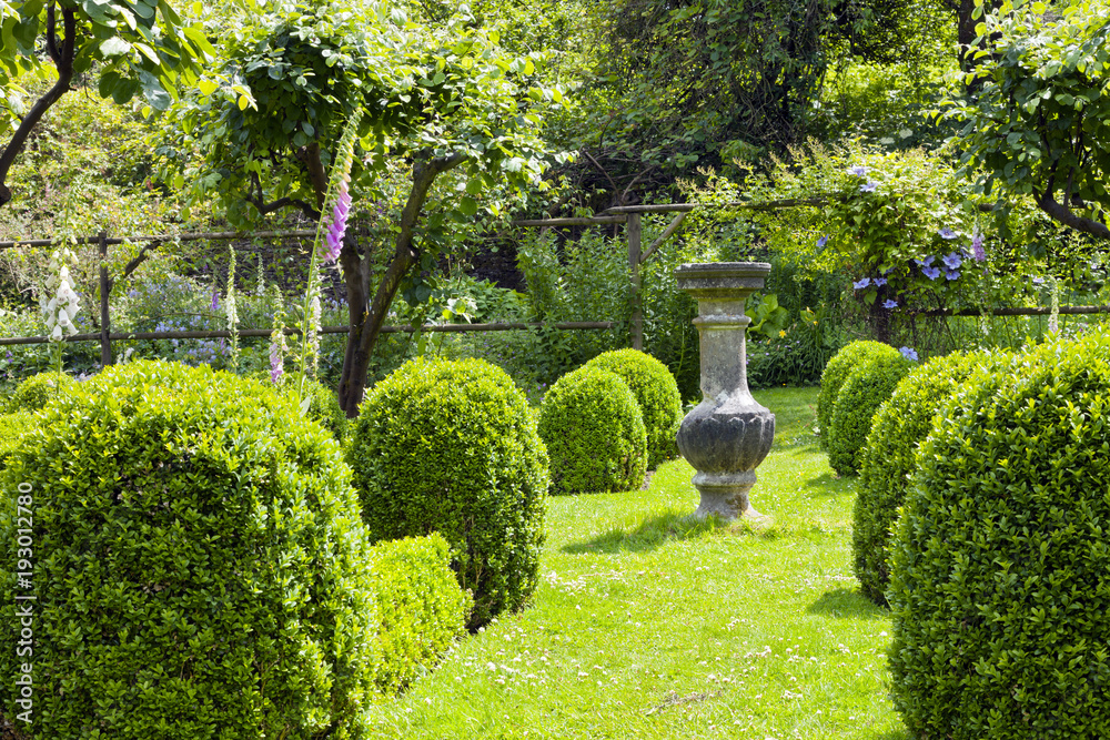 Stone ornamental vase on a grass path between buxus rounded topiary shrubs leading to a wooden arch with flowering clematis, in a cottage garden in rural England countryside .