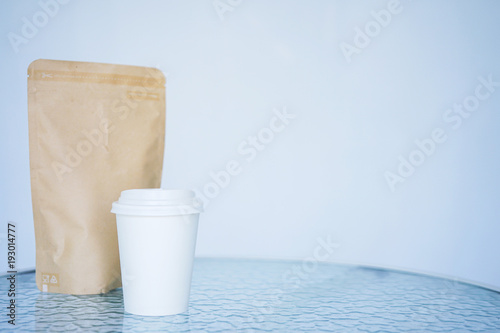 A white paper glass  one-time  for coffee a paper craft container is on the table. Background is blurred  white and blue. Place for the title on the glass.