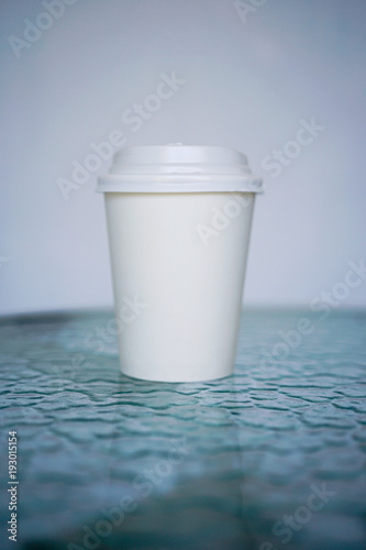 A white paper glass  one-time  for coffee is on the table. Background is blurred  white and blue. Place for the title on the glass. Side view with copy space