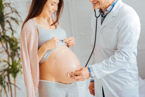 Healthy and strong. Cheerful doctor smiling while holding a stethoscope and listening to heartbeats of a little baby.