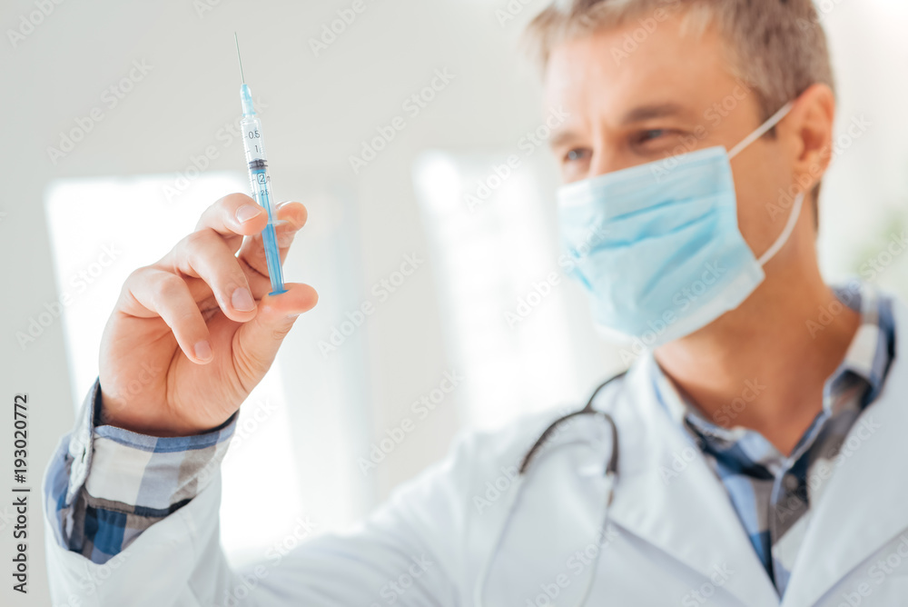 For injection. Careful joyful skilled medic in a gauze mask looking at the syringe holding it in right hand and dosing it. For injection. Careful joyful skilled medic in a gauze mask looking at the syringe holding it in right hand and dosing it.