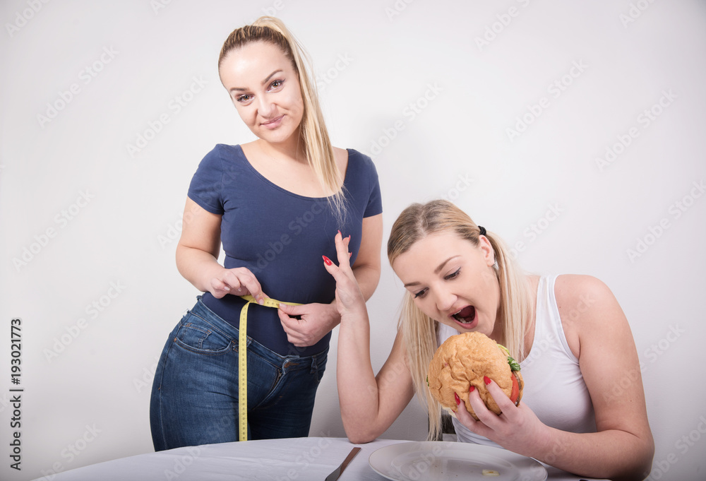 young blond woman eating while other one is measuring her waist young blond woman eating while other one is measuring her waist