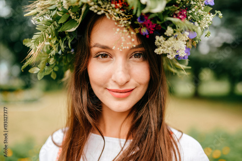 Closeup portrait of beautiful young girl in traditional Slavic dress with wreath of summer flowers. Ethno folk style cheerful female on abstract background in summer at floral feast. Expressive face. photo