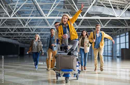Excited male and female people running to departure gate. One of them is wheeling baggage cart with woman sitting on the top