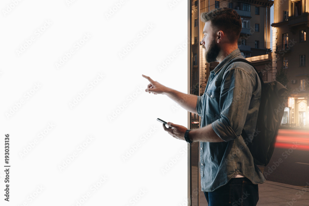 Summer night. Young hipster bearded man stands on city street and touches large glowing touchscreen while holding smartphone. Innovative technologies, digital display, urban navigation. Mock up. - obrazy, fototapety, plakaty 
