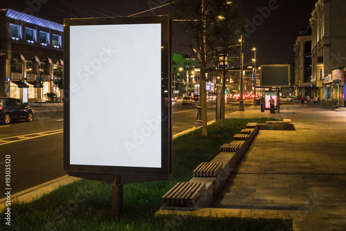 Vertical blank glowing billboard on night city street. In background buildings and road with cars. Mock up. Light box on street next to roadway. Space for logo, text, image, advertising, ad, blurb.