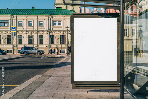 Vertical blank white billboard at bus stop on city street. In the background buildings and road. Mock up. Poster on street next to roadway. Sunny summer day.