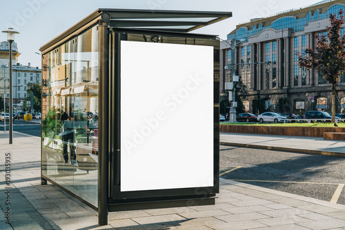 Vertical blank white billboard at bus stop on city street. In the background buildings and road. Mock up. Poster on street next to roadway. Sunny summer day. photo