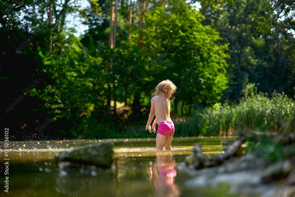 Adorable curly toddler girl wearing in pink shorts, playing at a river shore splashing water on sunny summer day Adorable curly toddler girl wearing in pink shorts, playing at a river shore splashing water on sunny summer day