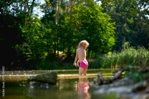 Adorable curly toddler girl wearing in pink shorts  playing at a river shore splashing water on sunny summer day