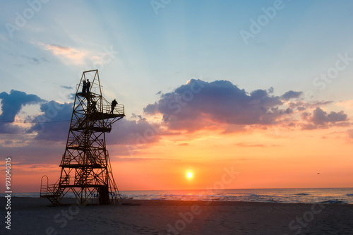 Silhouette of people on a rescue tower on sandy beach at sunset.