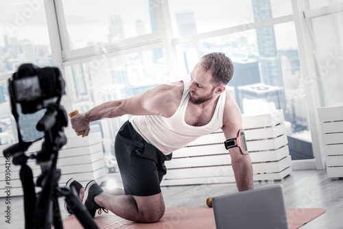 Heavy dumbbell. Concentrated focused male blogger using dumbbell while looking at it and exercising in front of camera