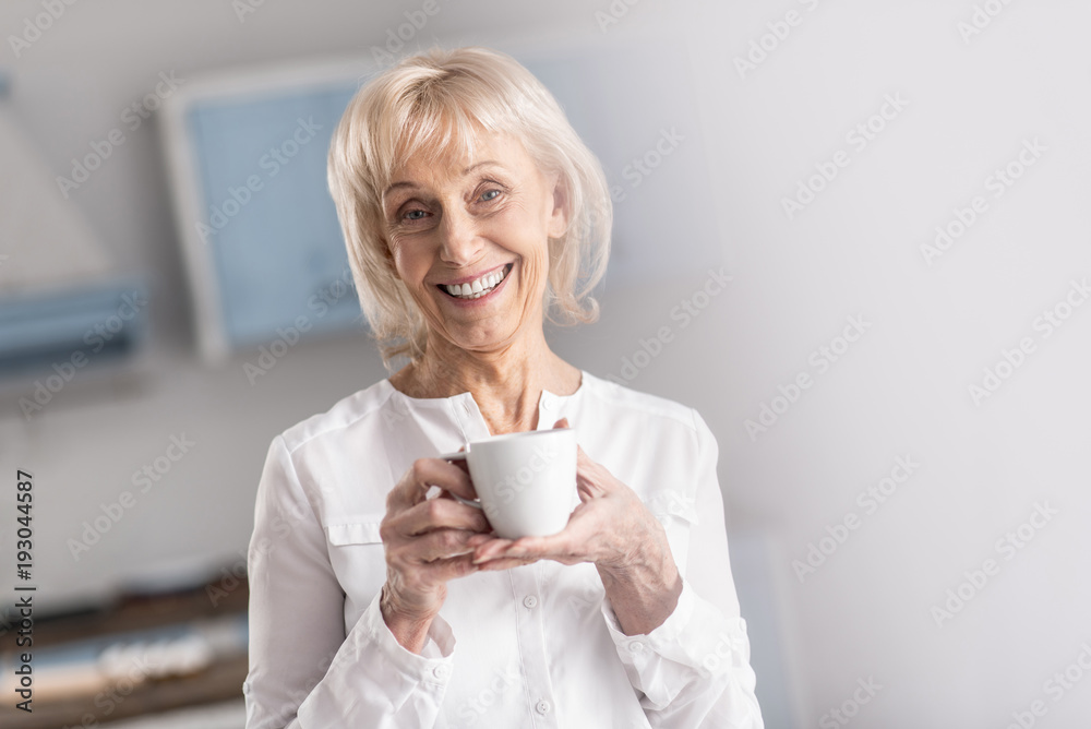 Morning coffee. Jolly merry mature woman looking at camera while carrying cup and smiling Morning coffee. Jolly merry mature woman looking at camera while carrying cup and smiling