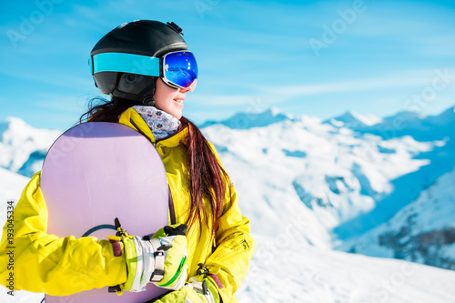 Picture of smiling woman in helmet and mask with snowboard on background of snowy hills