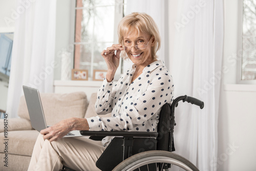 Stay aware. Disabled optimistic mature woman grinning in wheelchair while touching glasses and staring at camera